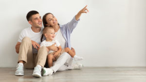 Happy Young Family With Infant Baby Sitting On Floor And Pointing Aside